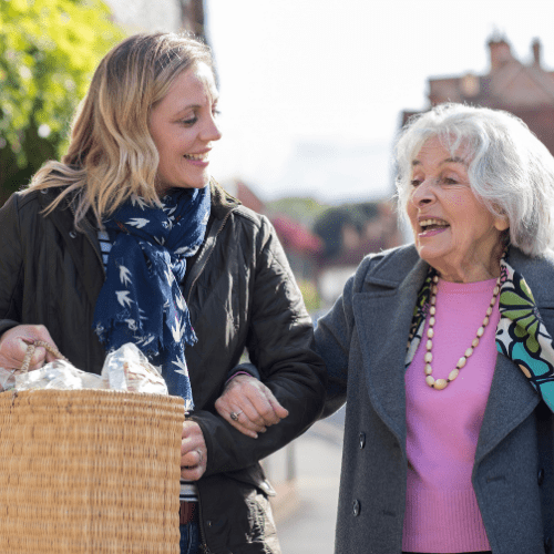 Two women walk together along a sunny street, smiling and chatting; the younger woman carries a wicker basket while guiding the elderly woman.